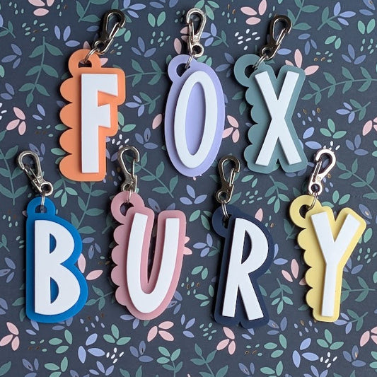 Six colorful letter-shaped keychains spelling 'FOX BURY' are arranged on a dark floral background. The letters are in orange, lavender, teal, blue, pink, and yellow, each with a silver metal clasp at the top.
